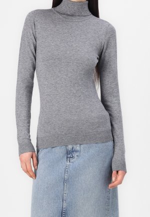 Femme portant un pull gris ajusté à col roulé associé à une jupe en denim bleu clair, debout devant un fond blanc uni.