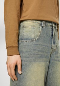 Person wearing light blue faded jeans and a brown long-sleeve shirt, with their left hand relaxed by their side.