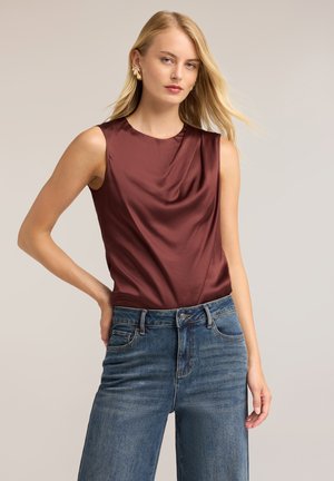 Femme aux longs cheveux blonds portant un haut en satin grenat sans manches et un jean bleu, debout avec une main sur la hanche devant un fond uni.