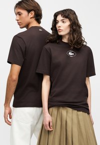 Un homme et une femme portant des t-shirts Lacoste marron foncé ; l'homme est de dos, la femme est de face avec une jupe plissée beige.