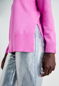Osoba o ciemnej karnacji, ubrana w jaskrawy różowy sweter z bocznym rozcięciem oraz metaliczne srebrne spodnie, stojąca na tle jednolitego jasnego tła.