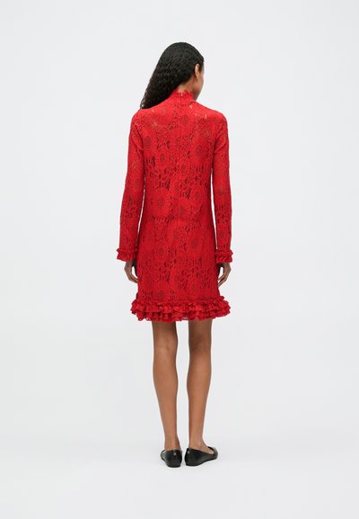 Piros csipkeblúz hosszú ujjakkal, fodros végű és virágmintás. Részletei között található a magas nyak és a texturált felület. Fekete lapos cipővel viselt.