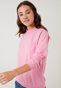 Felpa rosa con colletto alto, maniche lunghe e polsini a costine. Il tessuto sembra morbido e leggero, con un design minimalista.