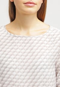 Blouse gris clair avec un motif hexagonal texturé, encolure ronde et léger éclat. Le tissu semble lisse avec un léger lustre.