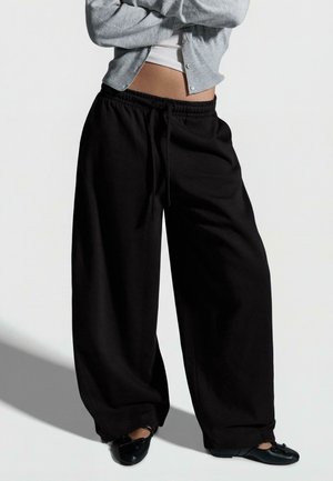 Persoană purtând pantaloni negri largi cu șnur, cardigan gri, top scurt alb și balerini negri, stând cu brațele încrucișate.