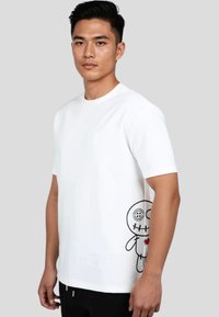 Hvid kortærmet T-shirt med rund hals. Har et sort tegneserie-design på den nederste side, der viser en syet karakter, der holder et hjerte.