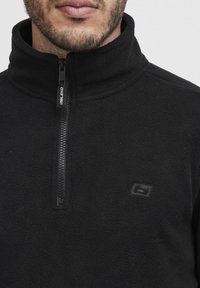 Sudadera de forro polar negra con cuello alto, cierre de cremallera y un pequeño logo en el lado izquierdo del pecho. Textura suave y diseño cálido.