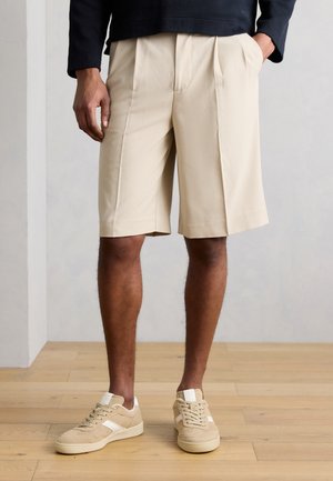 Mężczyzna model noszący beżowe spodenki z plisami i pasujące beżowe trampki, stojący na jasnej drewnianej podłodze z rękami w kieszeniach.