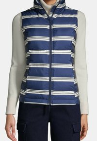 Marineblaue Steppweste mit horizontalen weißen Streifen, Reißverschluss und hohem Kragen, kombiniert mit einem cremefarbenen Rollkragenpullover und dunkler Hose.