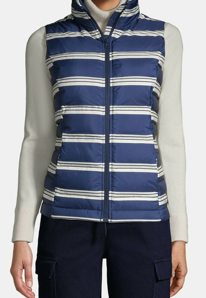 Marineblaue Steppweste mit horizontalen weißen Streifen, Reißverschluss und hohem Kragen, kombiniert mit einem cremefarbenen Rollkragenpullover und dunkler Hose.