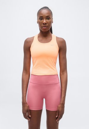 Atletische vrouw die recht vooruit kijkt, draagt een perzikkleurige mouwloze top en roze hoge taille fietsbroekje op een witte achtergrond.