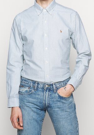 Mand iført blå- og hvidstribet skjorte med knapper, stoppet ned i blå denimjeans, med én hånd i lommen og synlig tatovering på håndleddet.