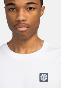 Camiseta blanca de algodón con un pequeño logo azul marino en el área del pecho izquierdo, cuello redondo y mangas cortas. Textura suave.