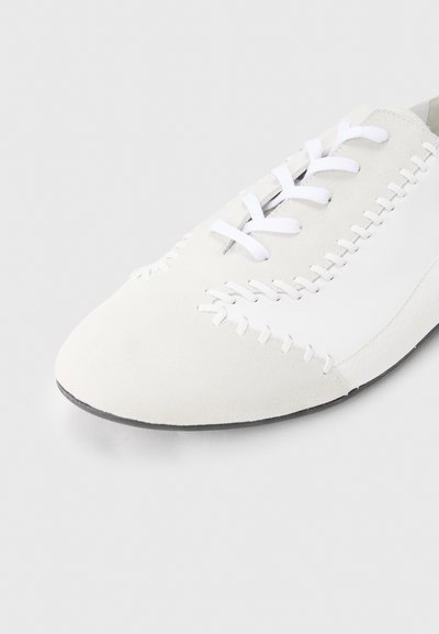 Άσπρο παπούτσι από σουέντ με μυτερή μύτη, διακοσμημένο με άσπρα κορδόνια και αντίθετες λεπτομέρειες ραφής κατά μήκος των άκρων. Μαύρη λαστιχένια σόλα.