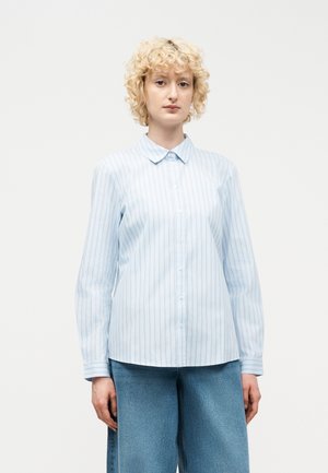 Personne aux cheveux blonds bouclés portant une chemise à rayures fines bleu clair boutonnée et un jean bleu à jambes larges, debout devant un fond uni.