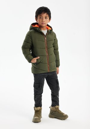 Giacca a vento verde con fodera arancione, dotata di chiusura con zip e tasche laterali. Abbinata a pantaloni cargo scuri e stivali color cammello.