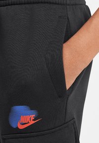 Pantalones de chándal de algodón negros con un bolsillo lateral, con un logo de Nike en azul y rojo impreso cerca del bolsillo para dar un acento. Textura suave.