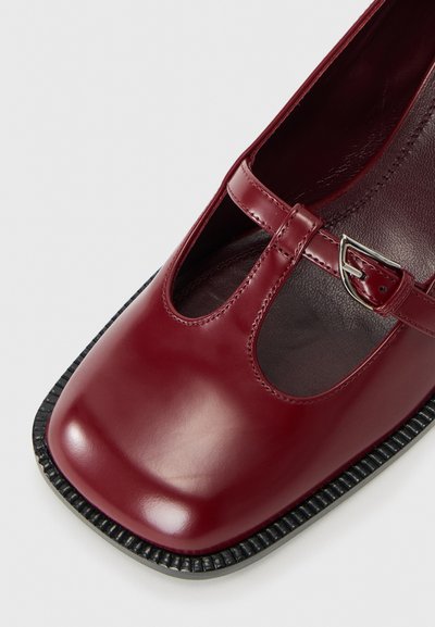 Chaussure Mary Jane en cuir bordeaux avec finition brillante, bout rond, lanière latérale avec boucle et semelle noire texturée.