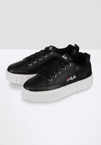 Zapatillas de cuero negro con suela de goma blanca plana, con cordones redondos y un logo en el lateral. Textura suave y diseño sencillo.