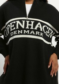 Pull en maille noir avec un col zippé mi-hauteur, présentant une bande horizontale blanche affichant "COPENHAGUE DANEMARK" en lettres épaisses.