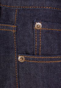 Tela de mezclilla oscura con costuras naranjas, que presenta dos bolsillos frontales y botones de herrajes en bronce. Primer plano del detalle de la textura y el diseño.