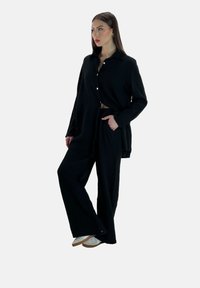 Conjunto negro hecho de tejido texturizado; camisa de botones de corte oversize con bolsillos frontales combinada con pantalones de pierna ancha y zapatillas blancas.
