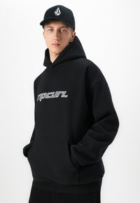 Mladý muž v čiernej mikine Ripcurl a čiernej čiapke s bielym logom, stojí s rukami v vreckách mikiny na bielom pozadí.