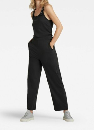 Femme portant une combinaison noire sans manches avec poches latérales et baskets grises, debout les mains dans les poches sur fond blanc.