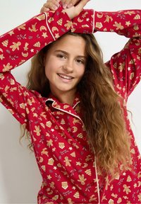 Rode pyjamashirt met feestelijke gouden patronen van peperkoekmannetjes, sterren en bomen. Beschikt over een kraag, knopen en afgewerkte randen. Zachte stof.