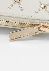 Portefeuille Versace Jeans en cuir synthétique gris clair avec des accents cloutés et une fermeture éclair ton or. Comprend une plaque métallique gravée "Versace Jeans".