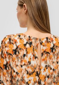 Plissé blouse in een multikleurig patroon met oranje, zwart en crème tinten. De achterkant heeft een kleine sleutelgatopening met een rits.