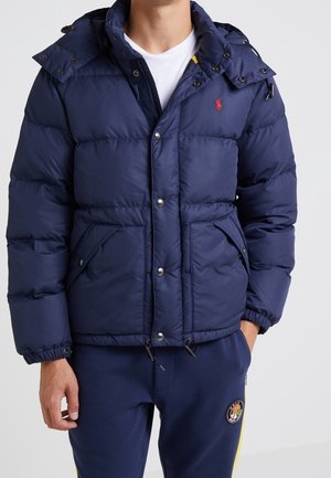 Man draagt een marineblauwe gewatteerde jas met capuchon over een wit T-shirt en bijpassende marineblauwe joggingbroek met logopatch op de linkerdij.