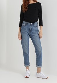 Leichtes schwarzes Langarmshirt, kombiniert mit hoch taillierten blauen Jeans, aufgerollten Ärmeln und weißen Sneakers mit orangen Akzenten.