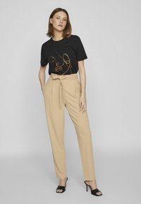 Zwarte t-shirt met een oranje line-art grafiek, gecombineerd met hooggewaisted beige broeken met een strikceintuur. Zwarte hoge hakken sandaal.