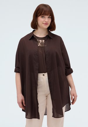 Vrouw met kort bruin haar draagt een loszittend donkerbruin overhemd met knopen over een kanten top en lichtbeige broek, staand tegen een effen achtergrond.