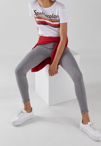 Jeans ajustados grises, camiseta blanca con rayas coloridas, suéter rojo atado a la cintura y zapatillas blancas en un modelo sentado contra un fondo blanco.