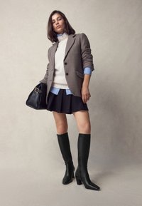 Svarta knähöga stövlar med klack, slätt läder, fyrkantig tå. Modell i hundtandsmönstrad kavaj, krämfärgad tröja, svart kjol, blå skjorta, håller en svart väska.