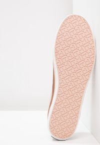 Semelle d’une basket rose clair avec un motif géométrique, positionnée à la verticale contre une surface blanche en angle.