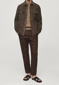 Uomo che indossa una giacca in suede marrone, un maglione beige a coste, pantaloni marrone scuro e sandali neri, in piedi con le mani nelle tasche.