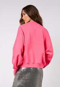 Jasně růžová bavlněná mikina s volným střihem, spadlými rameny a žebrovanými manžetami, spárovaná se stříbrnou flitrovou sukní.