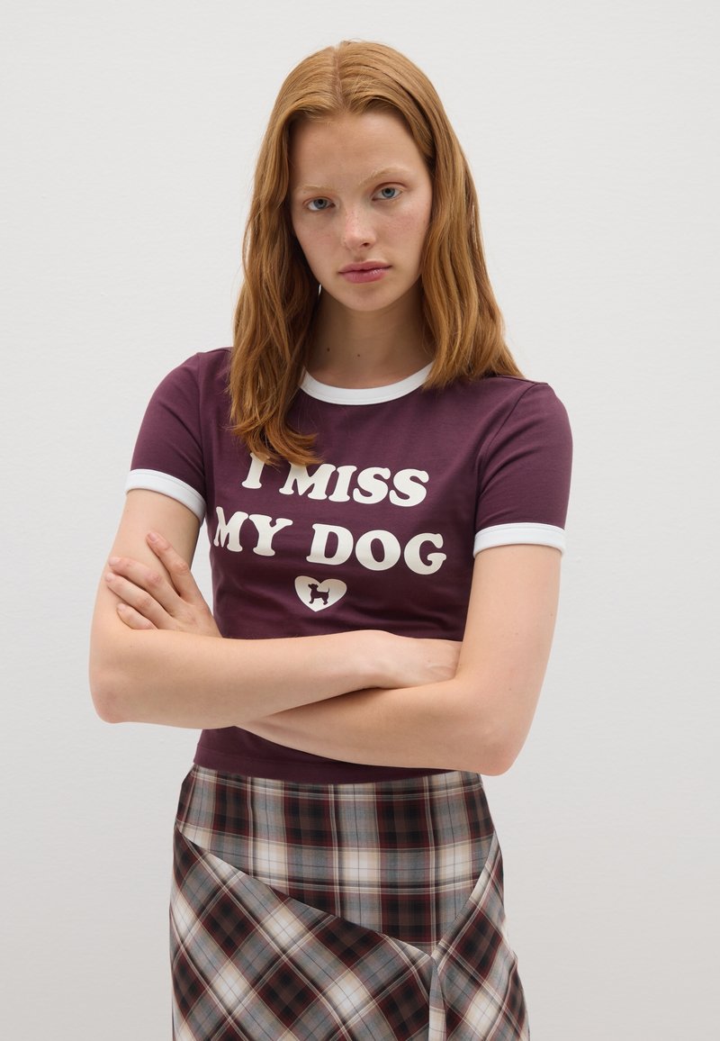 Una mujer joven con cabello rojo está de pie con los brazos cruzados, lleva una camiseta morada que dice "MI FALTA MI PERRO" y una falda de cuadros.