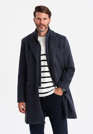 Abrigo texturizado azul marino con corte entallado y solapas con muesca, combinado con un suéter a rayas crema y azul marino. Jeans oscuros completan el conjunto.