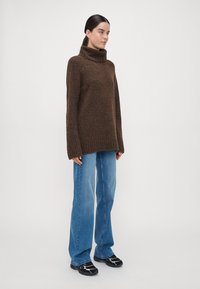 Übergroßer brauner Wollpullover mit hohem Kragen, gerippten Bündchen und Saum, kombiniert mit hellblauen, weitgeschnittenen Jeans und schwarzen Sneakers.