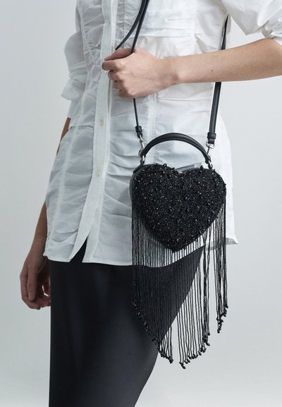 Άτομο ντυμένο με λευκό ρυχτό πουκάμισο και μαύρη φούστα, κρατώντας μια μαύρη τσάντα χειρός σε σχήμα καρδιάς με μακριές φούντες.