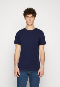 Tricou bleumarin cu mânecă scurtă, dintr-un material moale, cu un mic buzunar pe piept și guler rotund, asortat cu blugi albaștri.