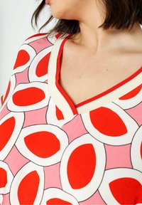 Kortærmet bluse med V-hals, mønstret med røde og hvide ovale former mod en lyserød baggrund. Accenteret med en rød kant.