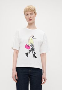 Hvit t-skjorte med ett grafisk illustrasjon av en figur i mønstrede bukser som bærer en rosa håndveske. Korte ermer, avslappet passform.