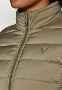 Veste rembourrée vert olive avec un col montant, dotée d'une fermeture éclair à l'avant et d'un patch logo triangulaire sur la poitrine. Texture lisse.