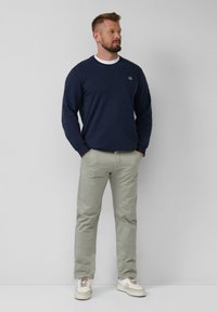 Sweat-shirt bleu marine avec col rond et logo, associé à un pantalon chino gris clair et des baskets crème avec des accents marron, se tenant debout de manière décontractée.