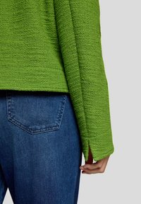 Grüner strukturierter Strickpullover mit lockerem Schnitt und Seitenschlitzen, kombiniert mit dunkelblauen Denim-Jeans mit schlichtem Design auf der Rückseite.